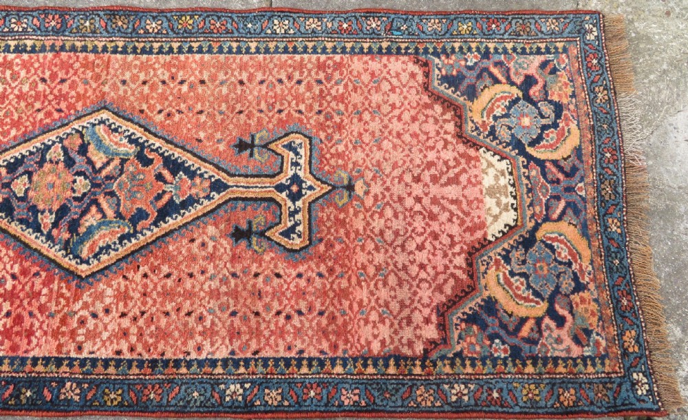 A long, narrow hand-knotted Kurdish runner rug featuring diamond medallions in navy and terracotta on a patterned rust-red field with decorative borders. View VI