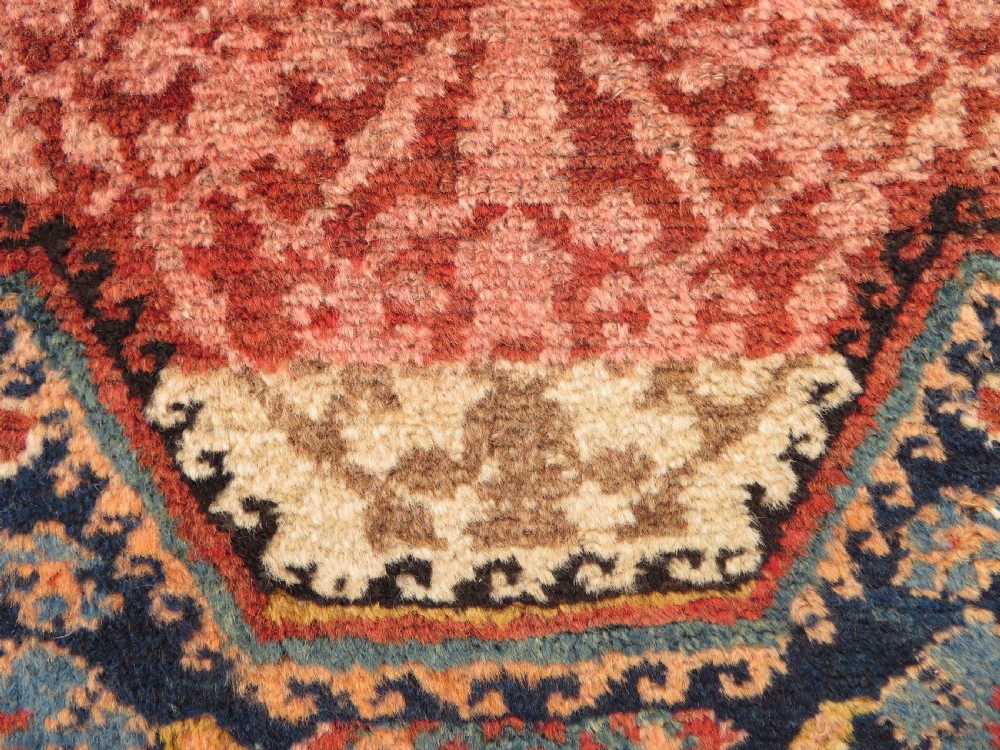 A long, narrow hand-knotted Kurdish runner rug featuring diamond medallions in navy and terracotta on a patterned rust-red field with decorative borders. View VII