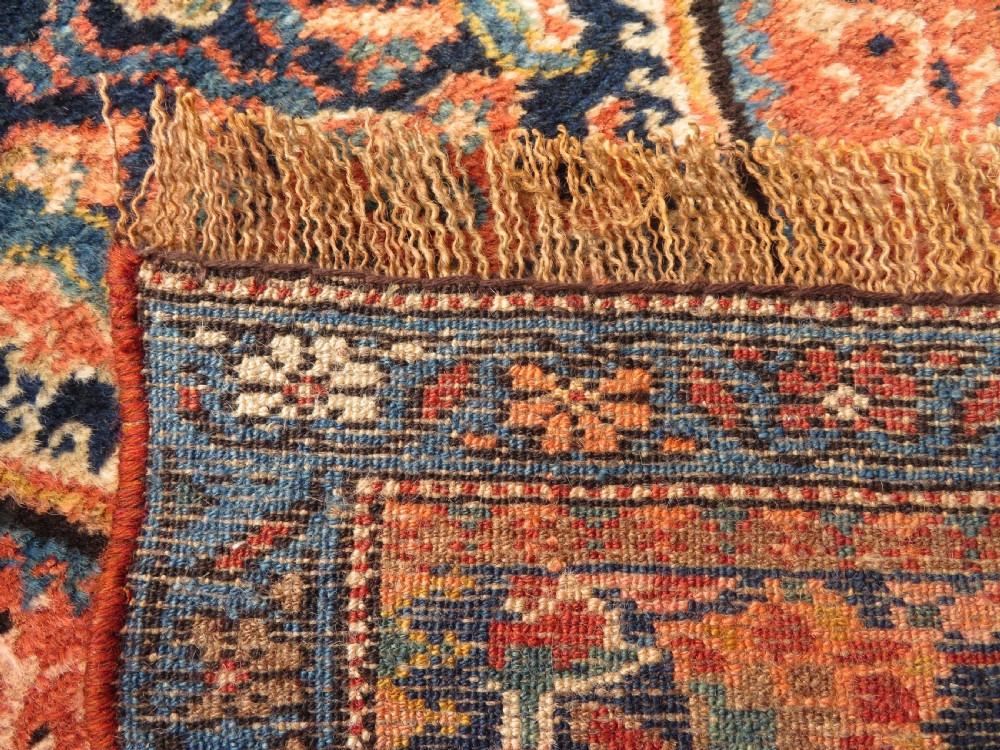 A long, narrow hand-knotted Kurdish runner rug featuring diamond medallions in navy and terracotta on a patterned rust-red field with decorative borders. View XV