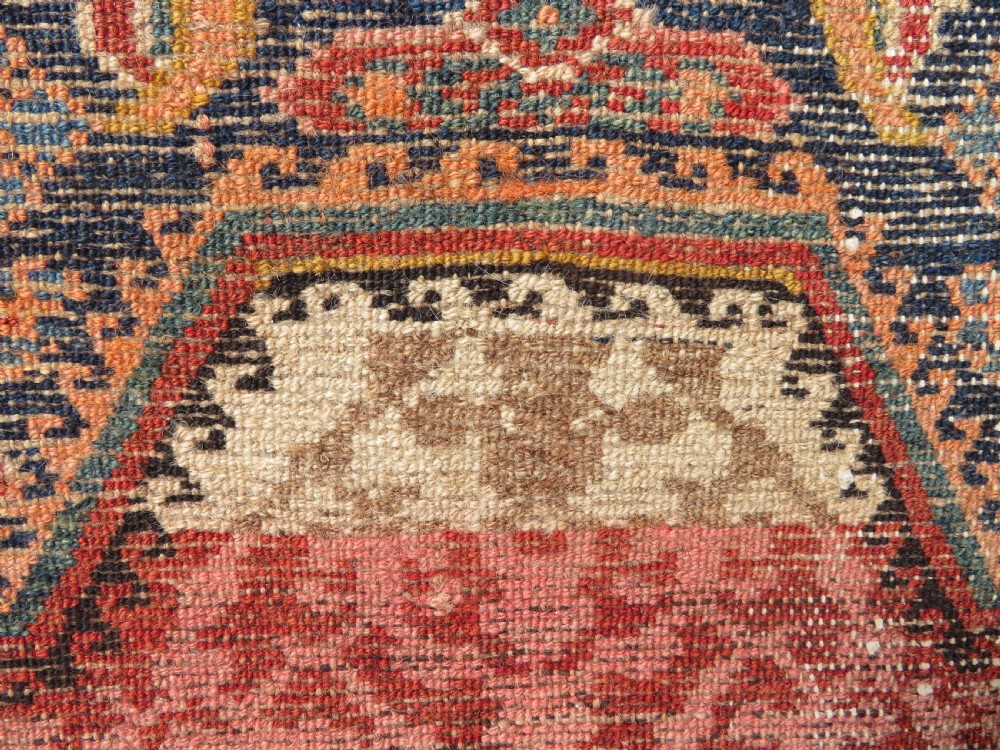 A long, narrow hand-knotted Kurdish runner rug featuring diamond medallions in navy and terracotta on a patterned rust-red field with decorative borders. View XXII