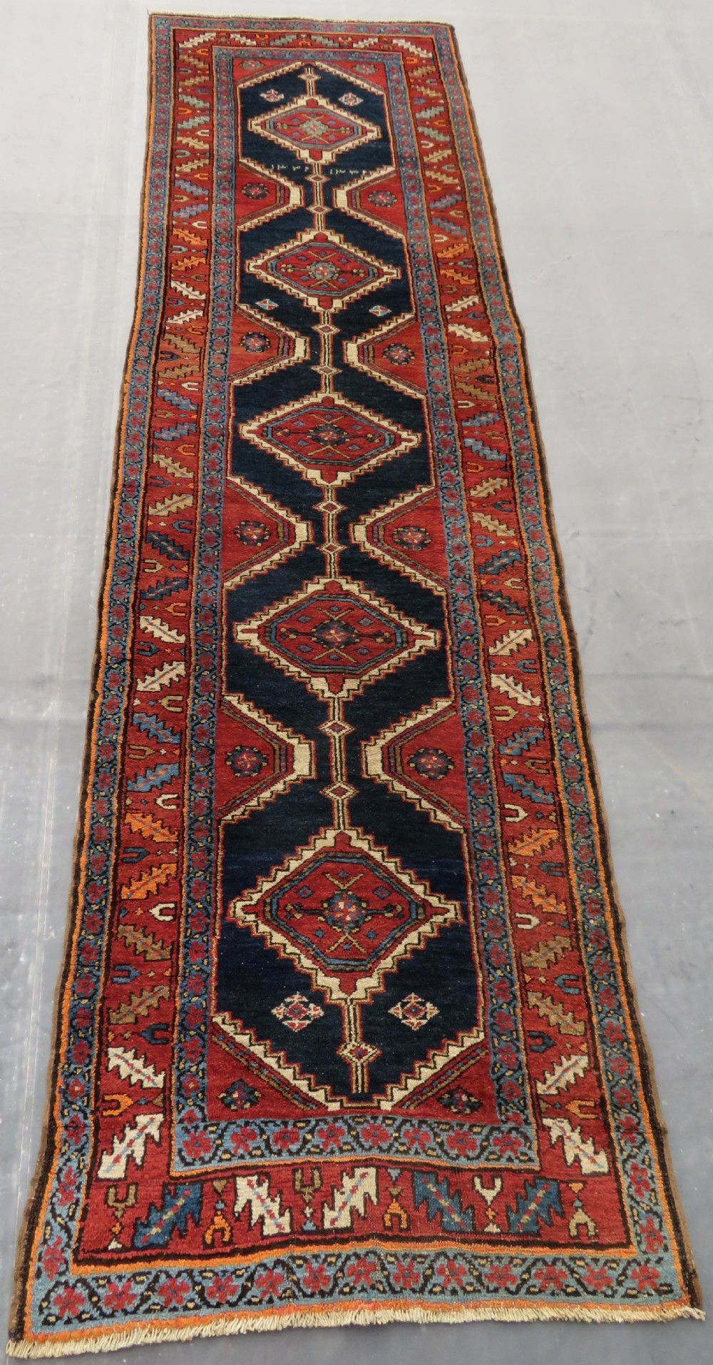 A long antique Persian Kelardasht runner rug featuring geometric diamond medallions on a dark navy blue central field framed by red and blue patterned borders.