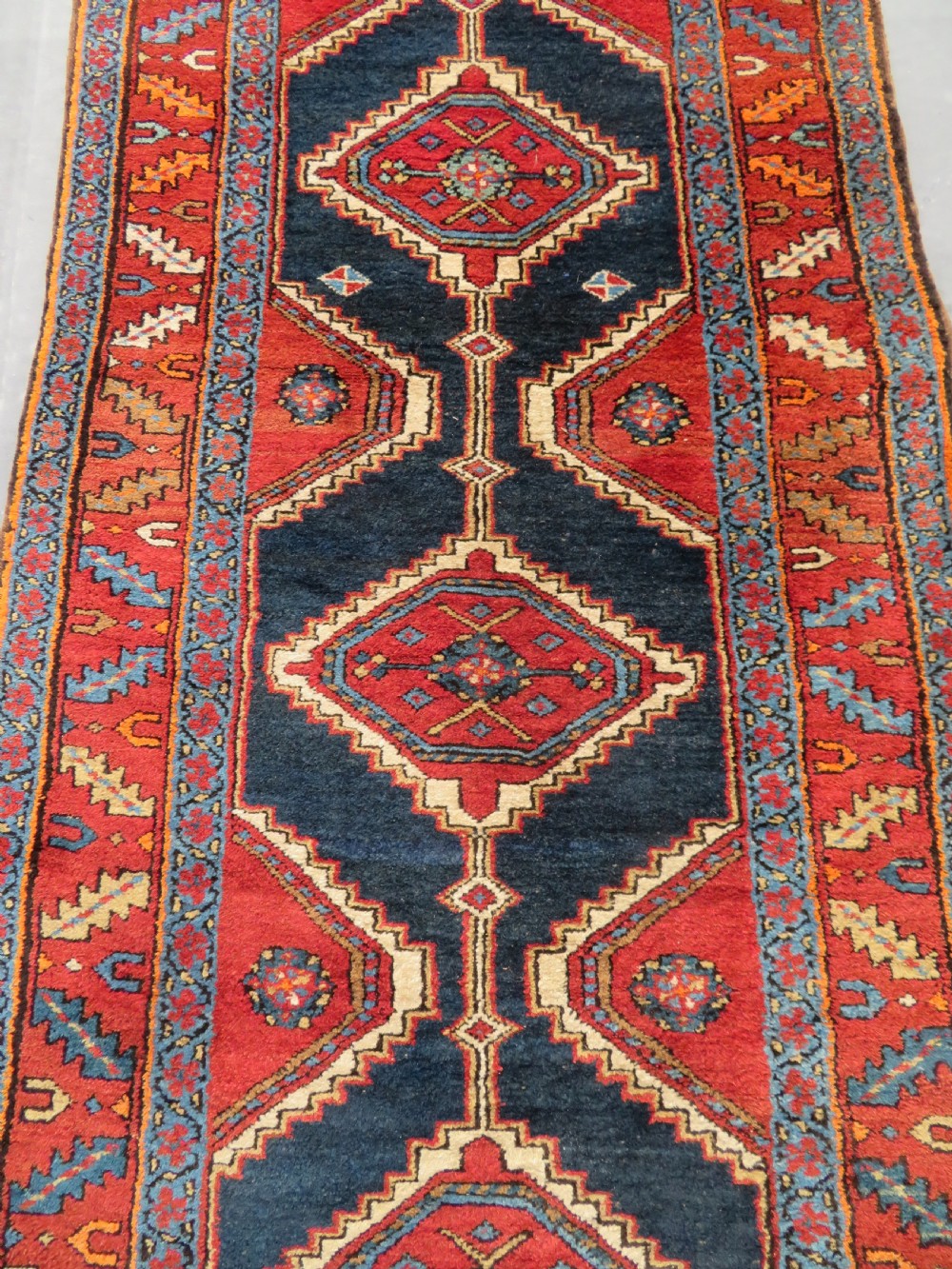 A long antique Persian Kelardasht runner rug featuring geometric diamond medallions on a dark navy blue central field framed by red and blue patterned borders. View V