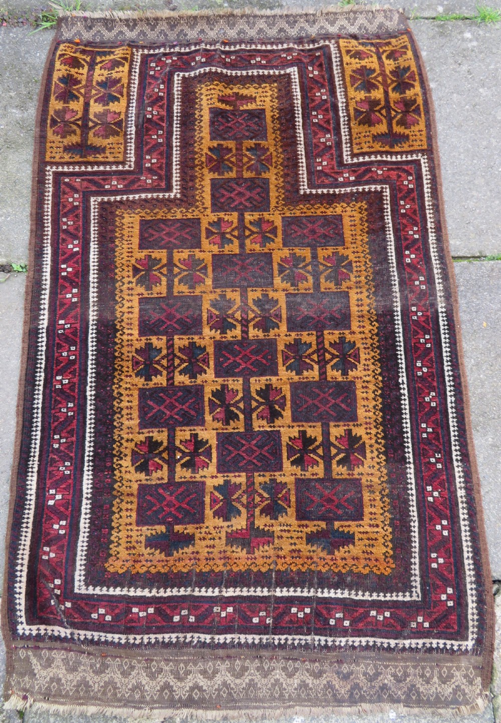An antique Baluch wool prayer rug featuring a stepped arched mihrab design on a camel-coloured field with deep red and navy geometric motifs.