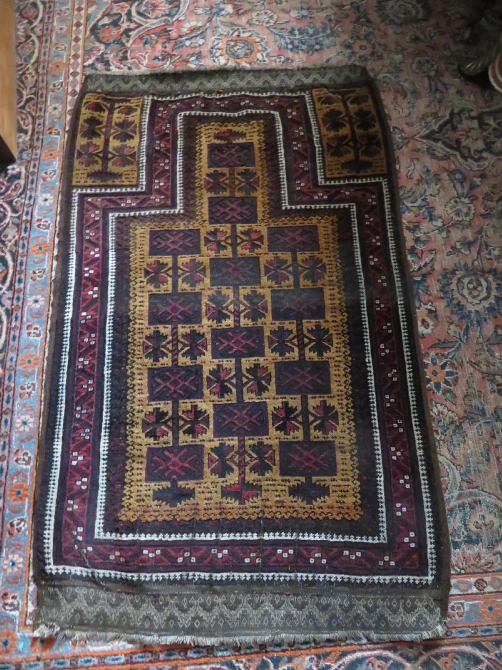 An antique Baluch wool prayer rug featuring a stepped arched mihrab design on a camel-coloured field with deep red and navy geometric motifs. View XX