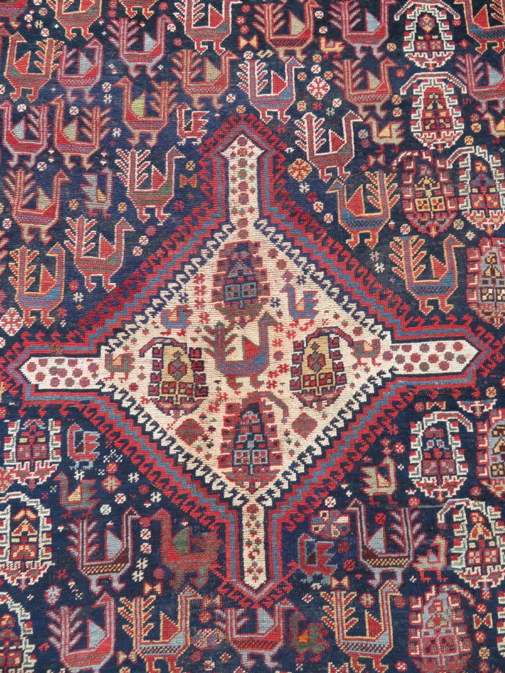 A late 19th-century Persian Qashqai hand-knotted wool rug featuring a navy blue field filled with stylized red peacock motifs, a central cream diamond medallion, and a wide red geometric border. View IV
