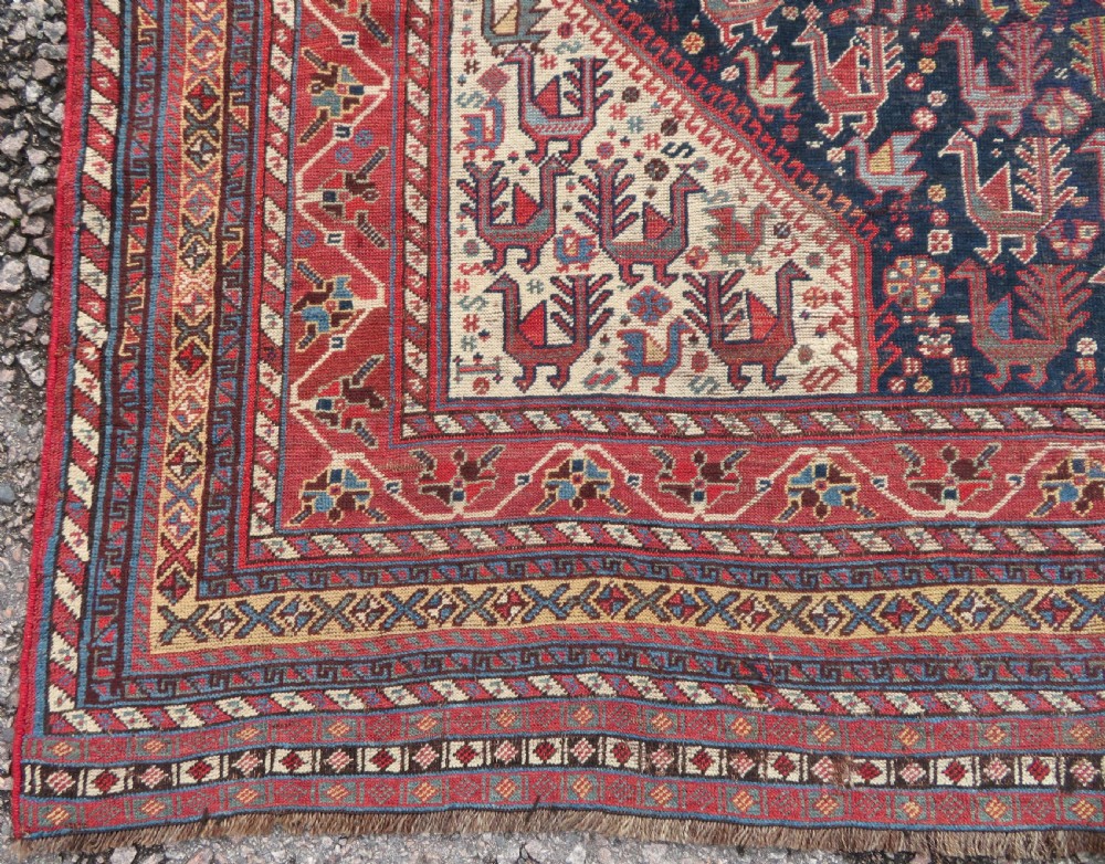 A late 19th-century Persian Qashqai hand-knotted wool rug featuring a navy blue field filled with stylized red peacock motifs, a central cream diamond medallion, and a wide red geometric border. View IX