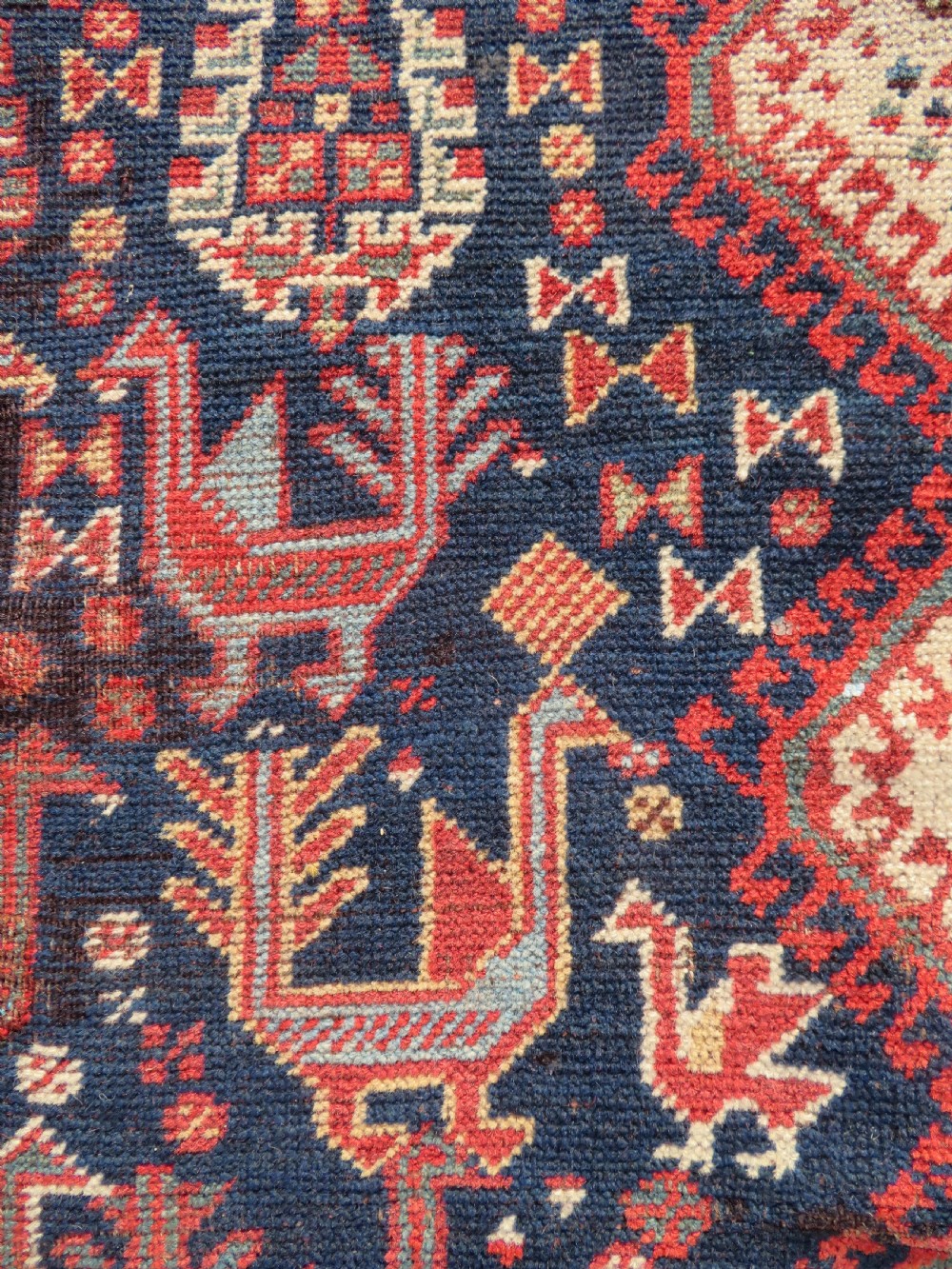A late 19th-century Persian Qashqai hand-knotted wool rug featuring a navy blue field filled with stylized red peacock motifs, a central cream diamond medallion, and a wide red geometric border. View XX