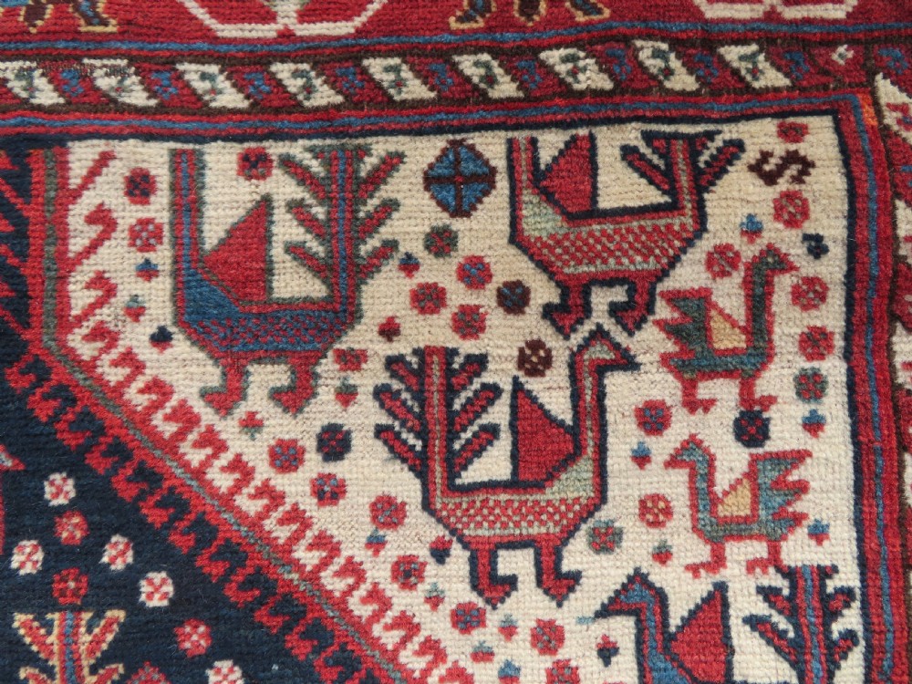 A late 19th-century Persian Qashqai hand-knotted wool rug featuring a navy blue field filled with stylized red peacock motifs, a central cream diamond medallion, and a wide red geometric border. View XXIV