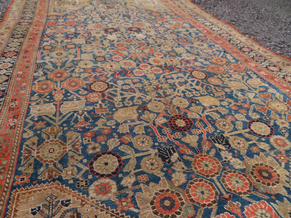 An antique Persian Qashqai Millefleurs wool rug featuring a central arch field filled with dense floral motifs in navy blue, terracotta, and ochre tones. View X