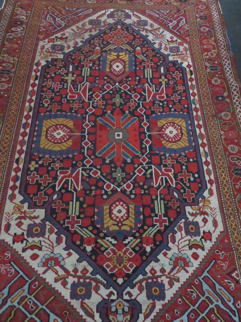 An antique hand-woven Persian Khamseh wool rug featuring a central geometric medallion with tribal bird motifs in red, navy blue, and ivory tones. View IV