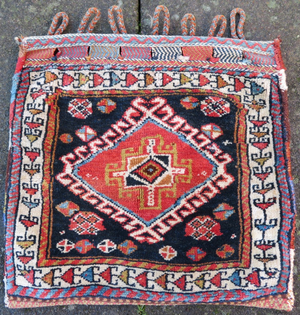 A square antique handwoven wool saddle bag face featuring a central red geometric diamond motif on a navy blue field with patterned cream borders and woven loops along the top edge.