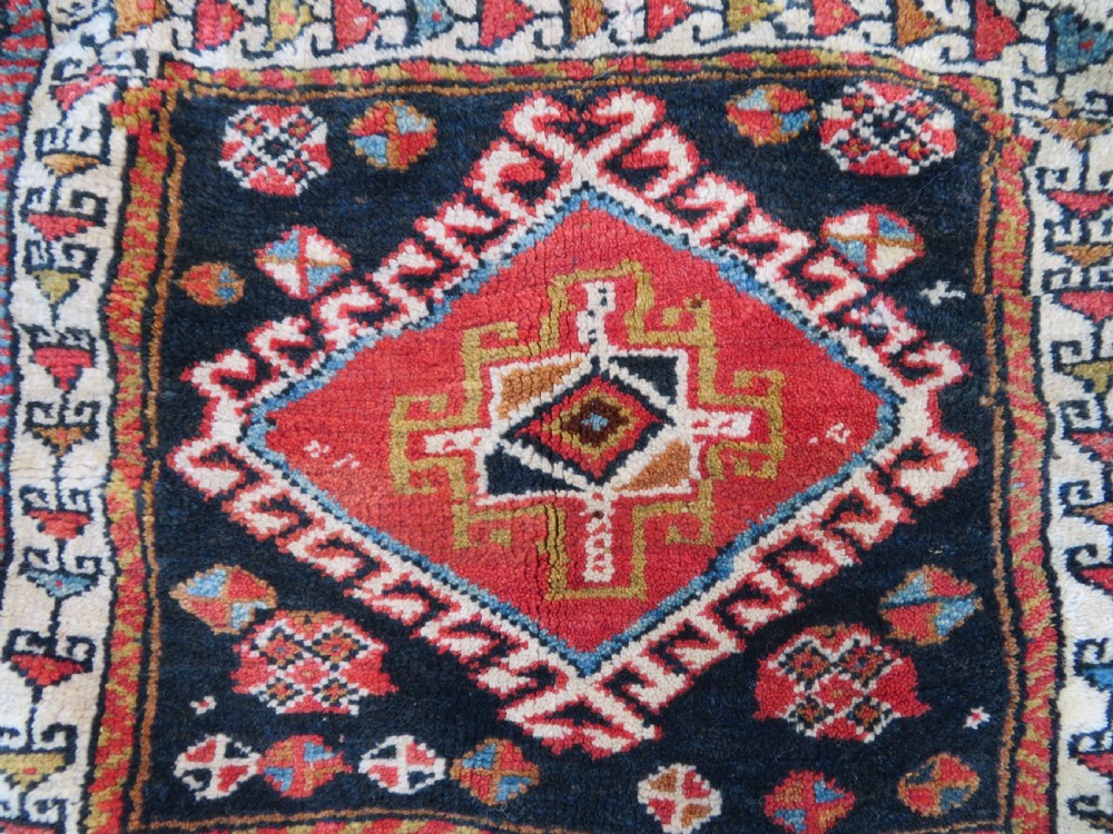 A square antique handwoven wool saddle bag face featuring a central red geometric diamond motif on a navy blue field with patterned cream borders and woven loops along the top edge. View II