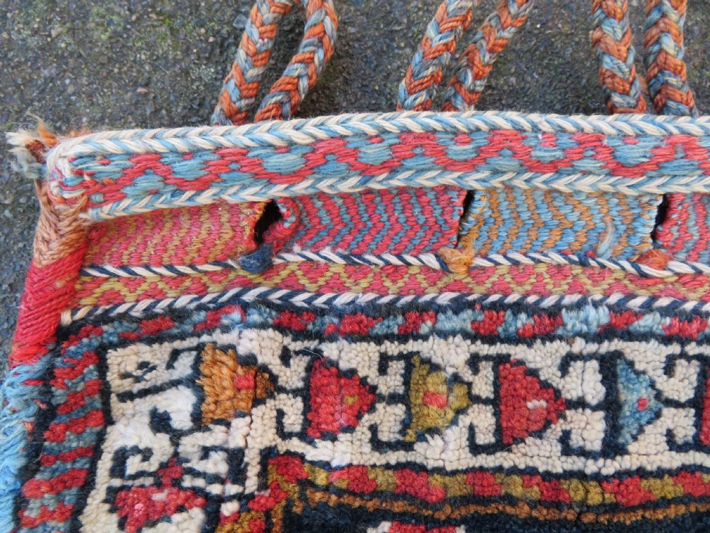 A square antique handwoven wool saddle bag face featuring a central red geometric diamond motif on a navy blue field with patterned cream borders and woven loops along the top edge. View III