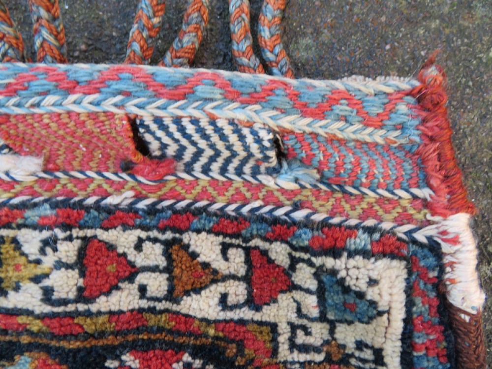 A square antique handwoven wool saddle bag face featuring a central red geometric diamond motif on a navy blue field with patterned cream borders and woven loops along the top edge. View IV