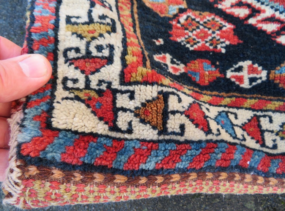 A square antique handwoven wool saddle bag face featuring a central red geometric diamond motif on a navy blue field with patterned cream borders and woven loops along the top edge. View V