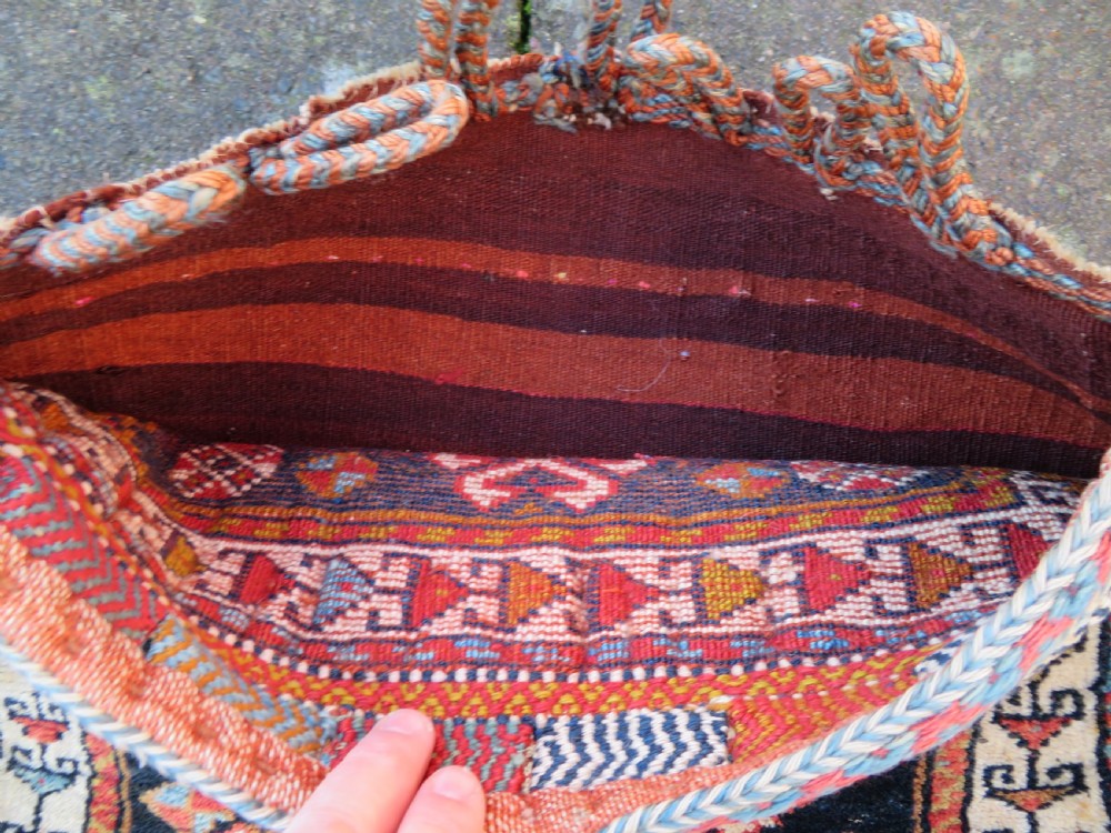 A square antique handwoven wool saddle bag face featuring a central red geometric diamond motif on a navy blue field with patterned cream borders and woven loops along the top edge. View VI