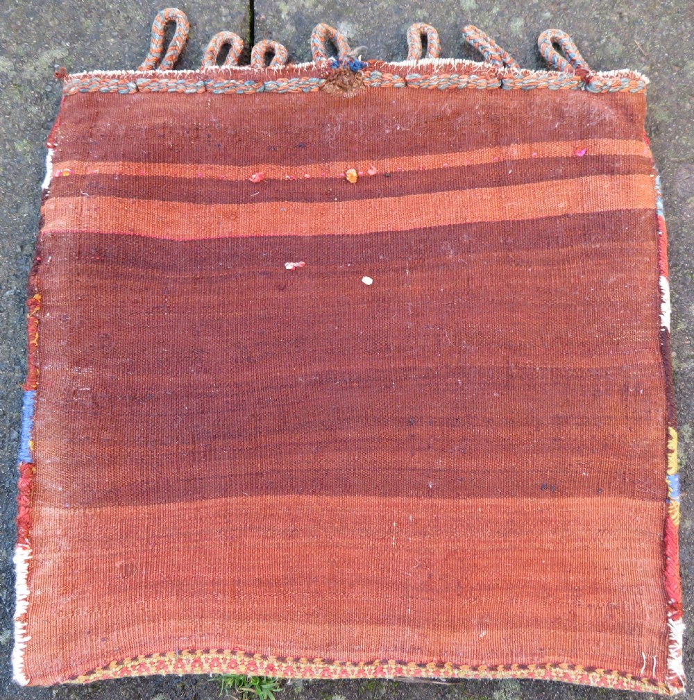 A square antique handwoven wool saddle bag face featuring a central red geometric diamond motif on a navy blue field with patterned cream borders and woven loops along the top edge. View VII