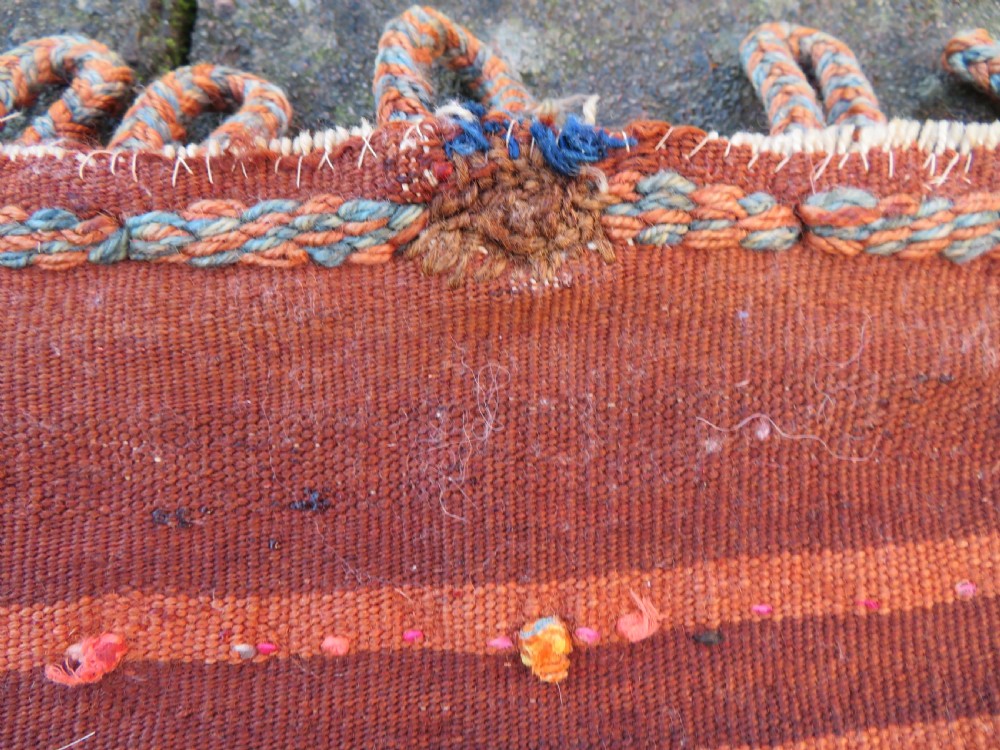A square antique handwoven wool saddle bag face featuring a central red geometric diamond motif on a navy blue field with patterned cream borders and woven loops along the top edge. View VIII