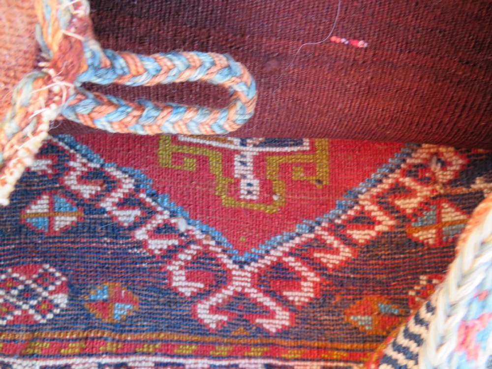 A square antique handwoven wool saddle bag face featuring a central red geometric diamond motif on a navy blue field with patterned cream borders and woven loops along the top edge. View X