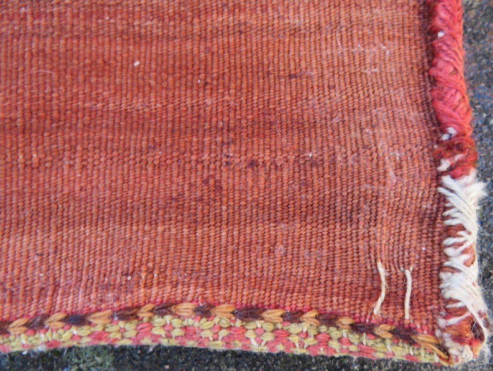 A square antique handwoven wool saddle bag face featuring a central red geometric diamond motif on a navy blue field with patterned cream borders and woven loops along the top edge. View XI