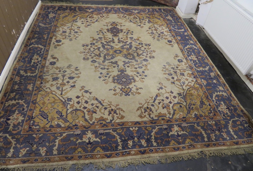An antique hand-knotted wool area carpet featuring an ivory central field, detailed central medallion, corner spandrels, and a broad indigo floral border.
