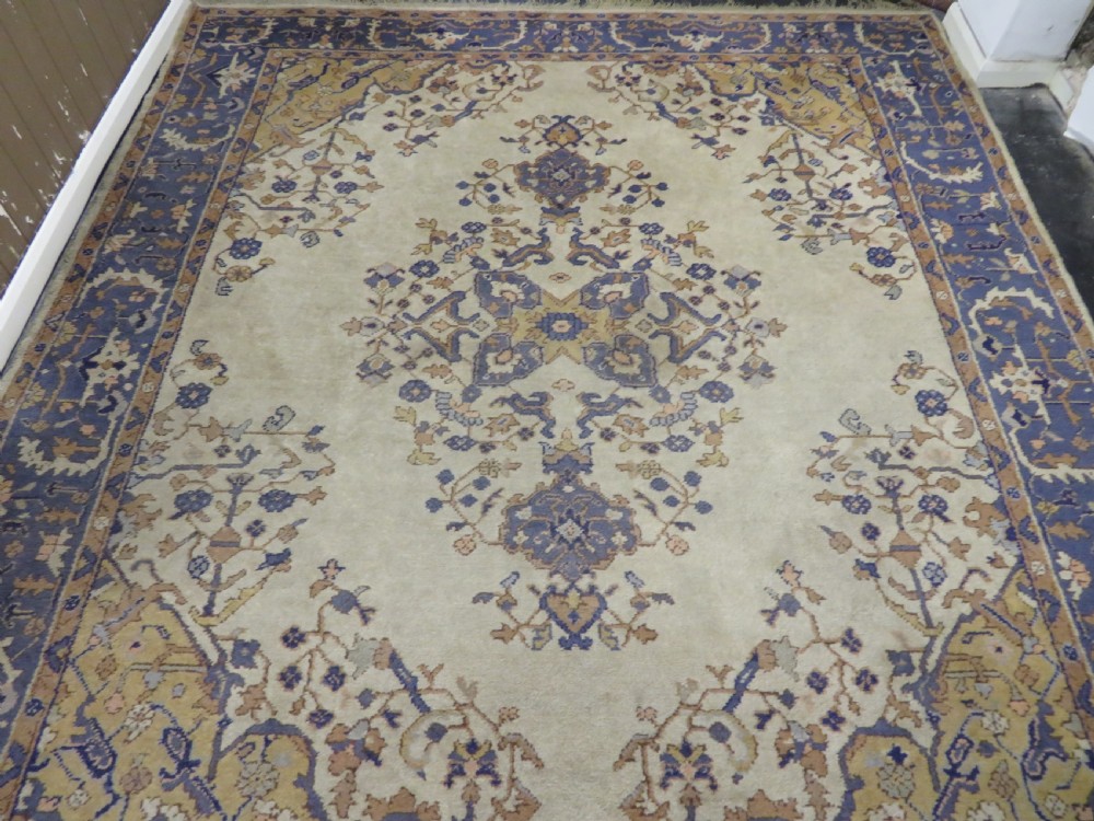 An antique hand-knotted wool area carpet featuring an ivory central field, detailed central medallion, corner spandrels, and a broad indigo floral border. View V