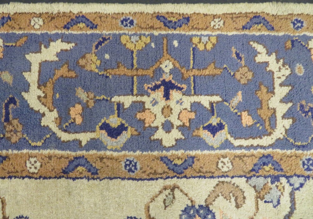 An antique hand-knotted wool area carpet featuring an ivory central field, detailed central medallion, corner spandrels, and a broad indigo floral border. View IX