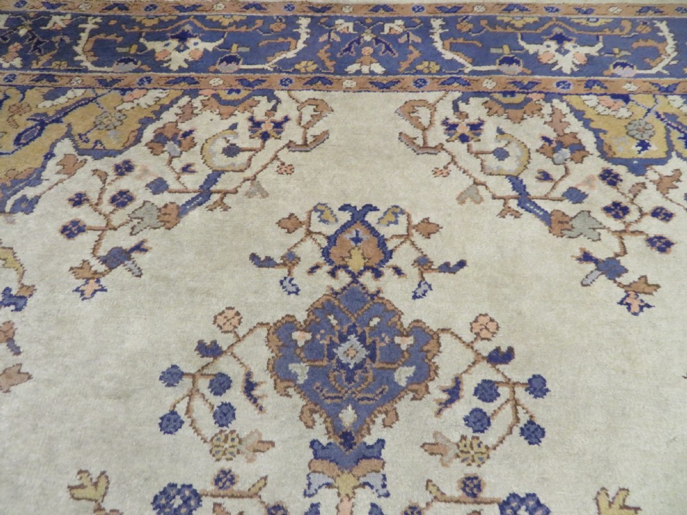 An antique hand-knotted wool area carpet featuring an ivory central field, detailed central medallion, corner spandrels, and a broad indigo floral border. View XVI