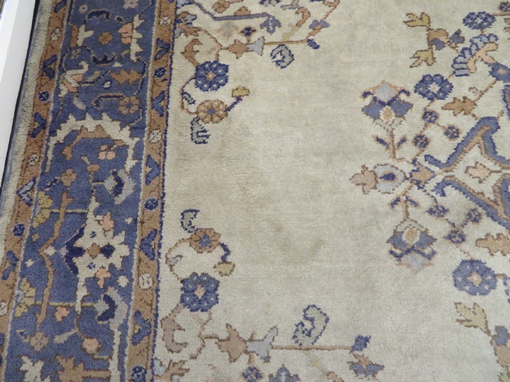 An antique hand-knotted wool area carpet featuring an ivory central field, detailed central medallion, corner spandrels, and a broad indigo floral border. View XVII