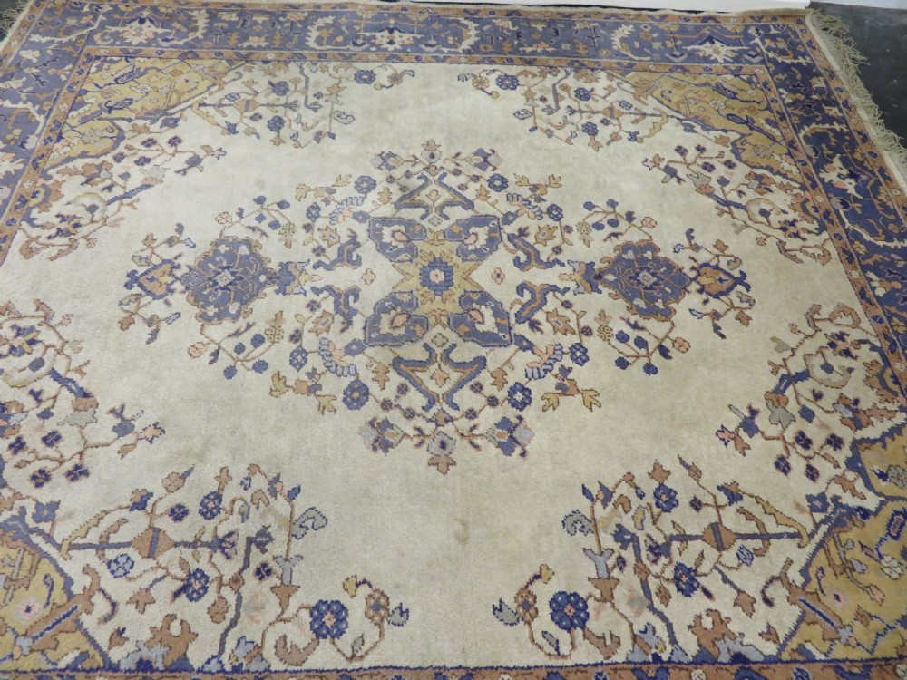 An antique hand-knotted wool area carpet featuring an ivory central field, detailed central medallion, corner spandrels, and a broad indigo floral border. View XIX