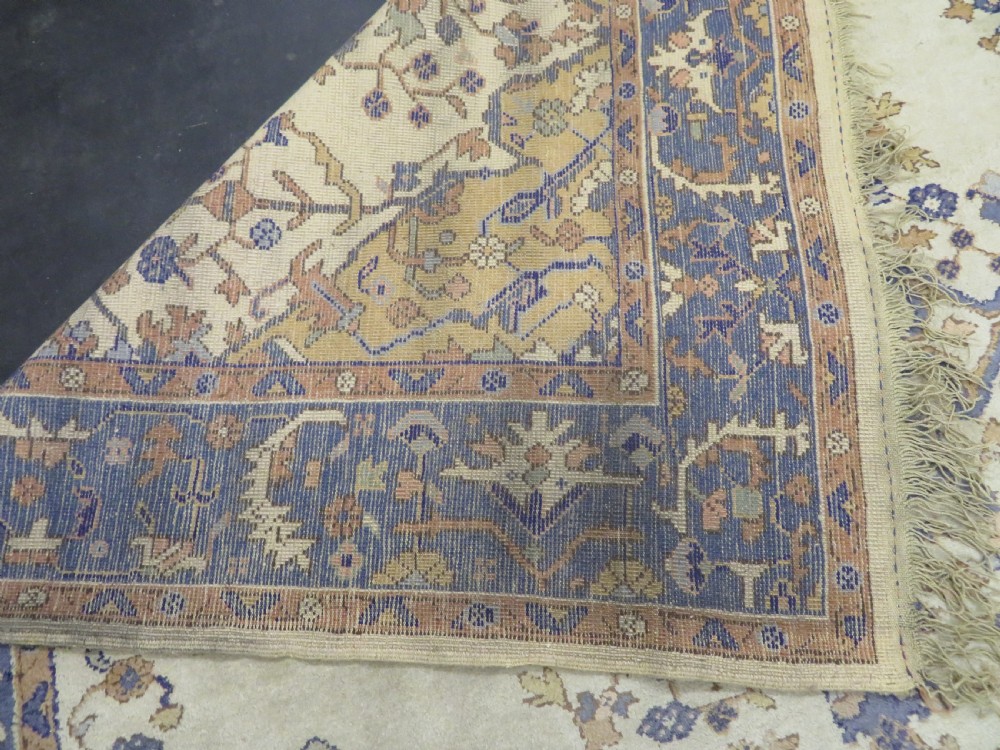 An antique hand-knotted wool area carpet featuring an ivory central field, detailed central medallion, corner spandrels, and a broad indigo floral border. View XXVI