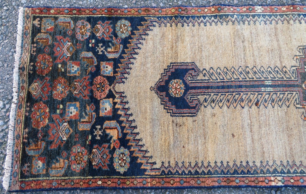 A long, narrow hand-knotted antique Persian Kurdish wool runner rug with geometric central column motifs on a camel field bordered in dark navy blue. View IV