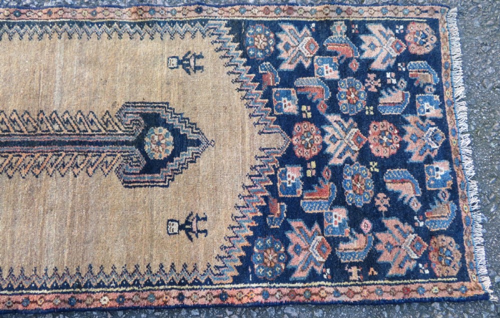 A long, narrow hand-knotted antique Persian Kurdish wool runner rug with geometric central column motifs on a camel field bordered in dark navy blue. View V