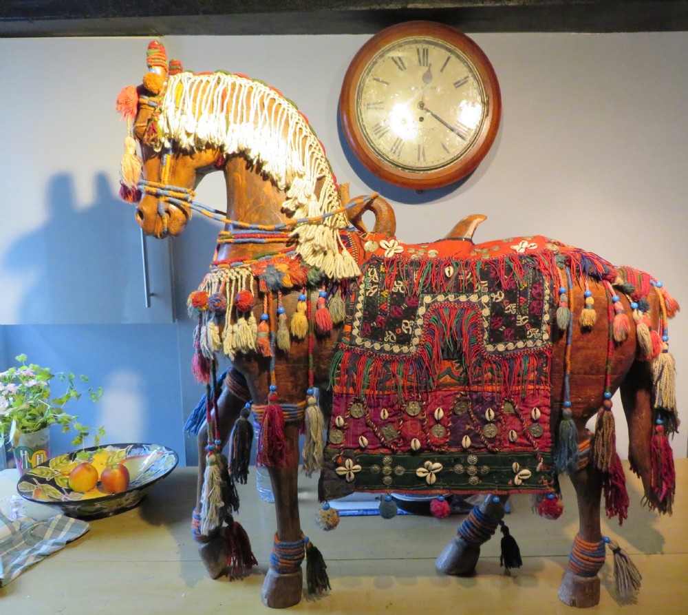 A large carved hardwood model of a standing horse decorated with detailed woven textiles, tassels, and cowrie shells.
