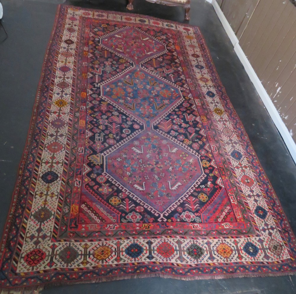 An antique hand-knotted Qashqai wool rug featuring three diamond central medallions with tribal peacock motifs in red, blue, and magenta tones.