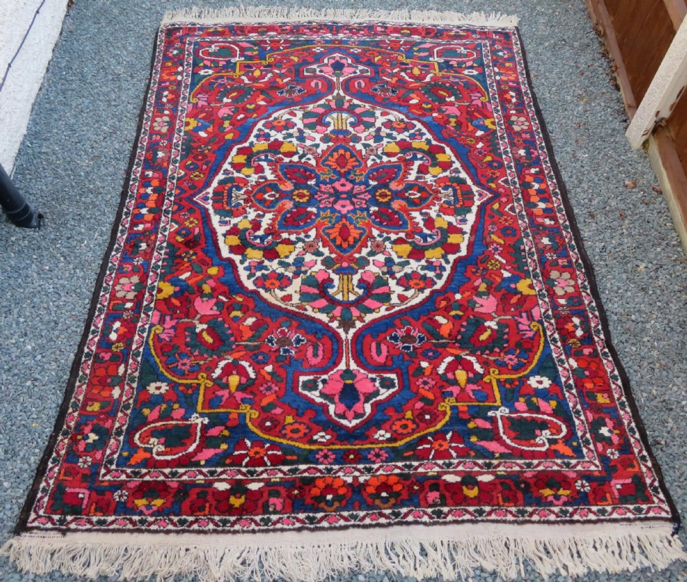 An antique Bakhtiari wool rug featuring a cream and pink central floral medallion framed by red and navy blue fields with complex botanical borders.