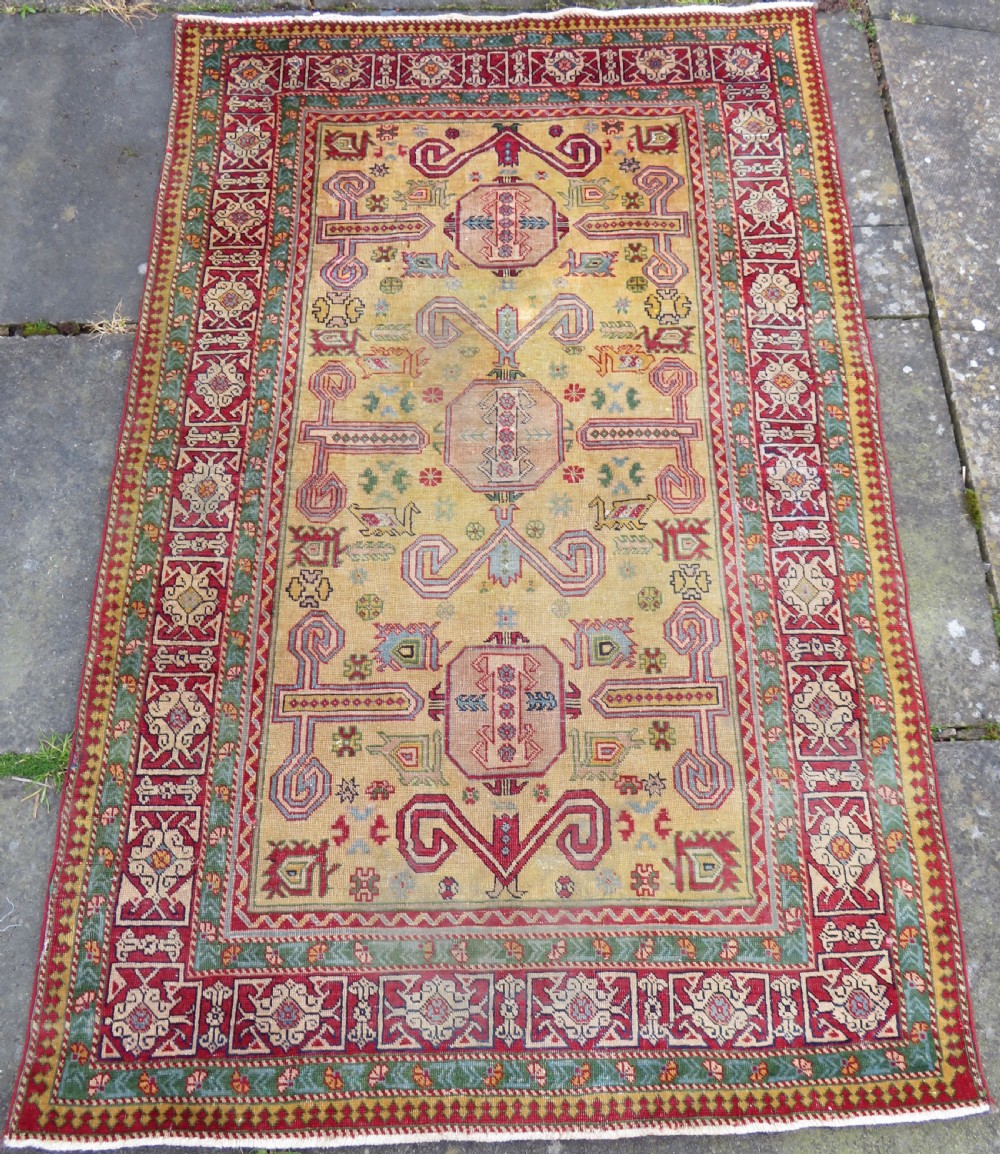 An antique hand-knotted Caucasian rug featuring a distressed yellow central field patterned with geometric ram horn motifs framed by red and green decorative borders.