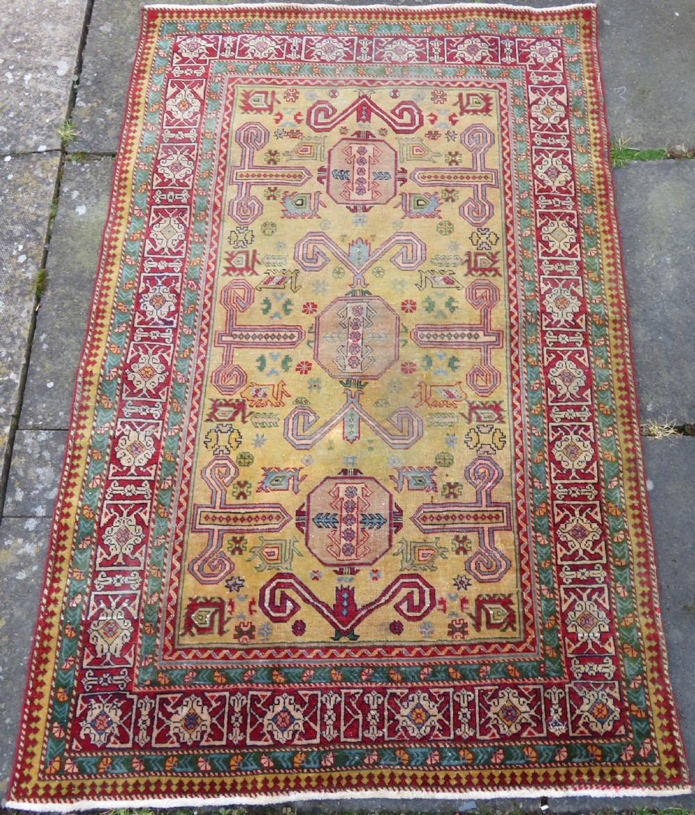An antique hand-knotted Caucasian rug featuring a distressed yellow central field patterned with geometric ram horn motifs framed by red and green decorative borders. View II