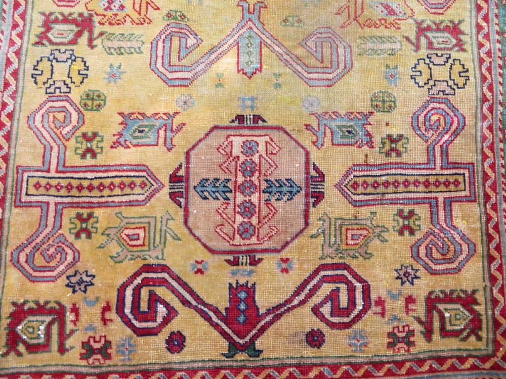 An antique hand-knotted Caucasian rug featuring a distressed yellow central field patterned with geometric ram horn motifs framed by red and green decorative borders. View III