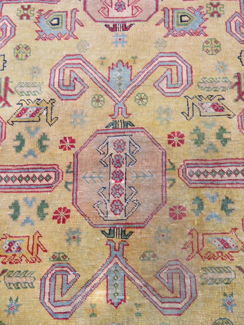An antique hand-knotted Caucasian rug featuring a distressed yellow central field patterned with geometric ram horn motifs framed by red and green decorative borders. View IV