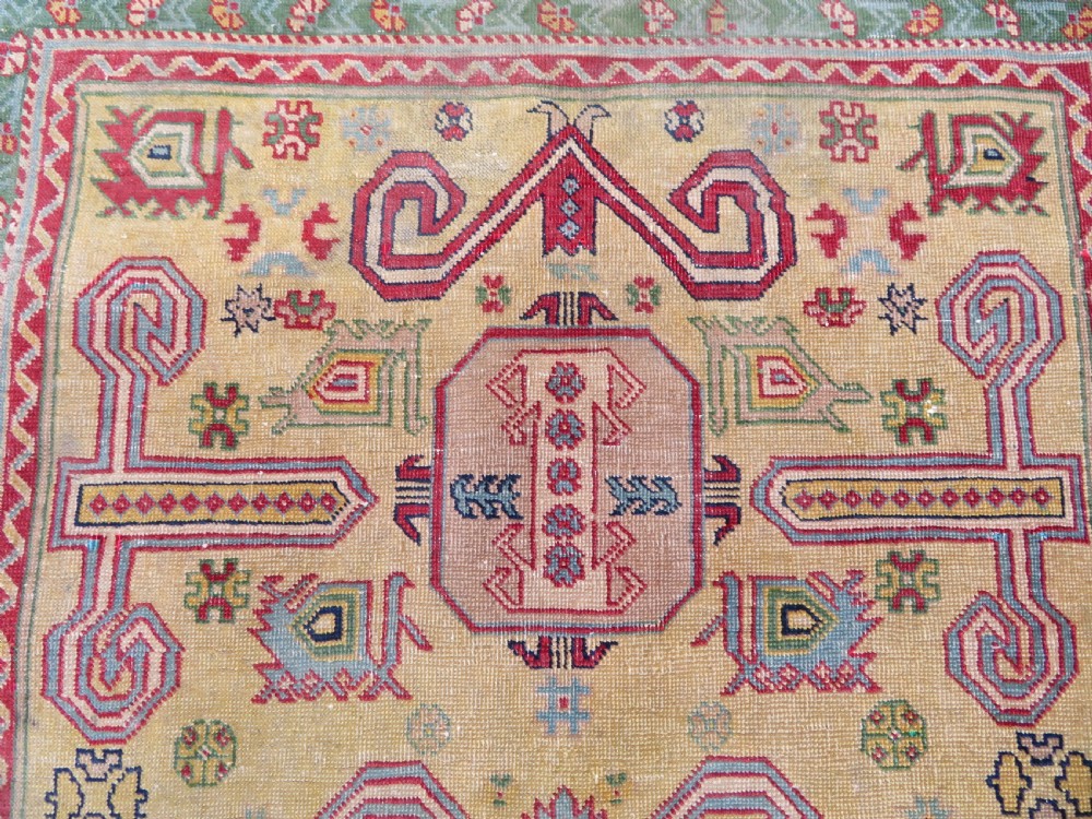 An antique hand-knotted Caucasian rug featuring a distressed yellow central field patterned with geometric ram horn motifs framed by red and green decorative borders. View V