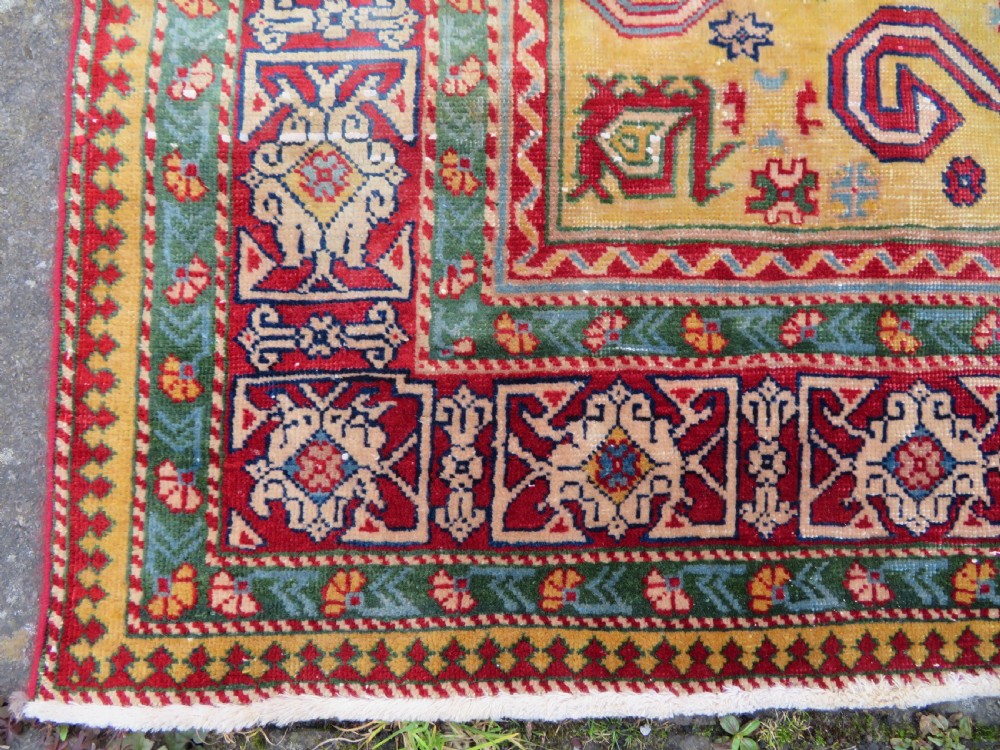 An antique hand-knotted Caucasian rug featuring a distressed yellow central field patterned with geometric ram horn motifs framed by red and green decorative borders. View VII