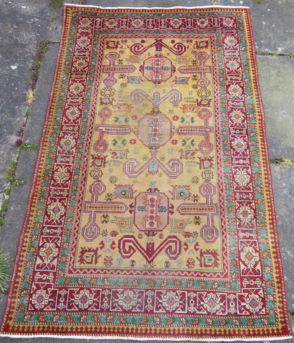 An antique hand-knotted Caucasian rug featuring a distressed yellow central field patterned with geometric ram horn motifs framed by red and green decorative borders. View IX