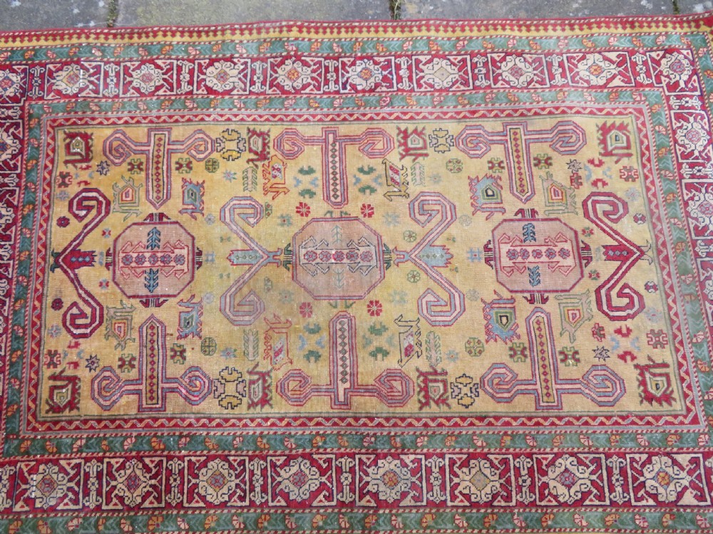 An antique hand-knotted Caucasian rug featuring a distressed yellow central field patterned with geometric ram horn motifs framed by red and green decorative borders. View X