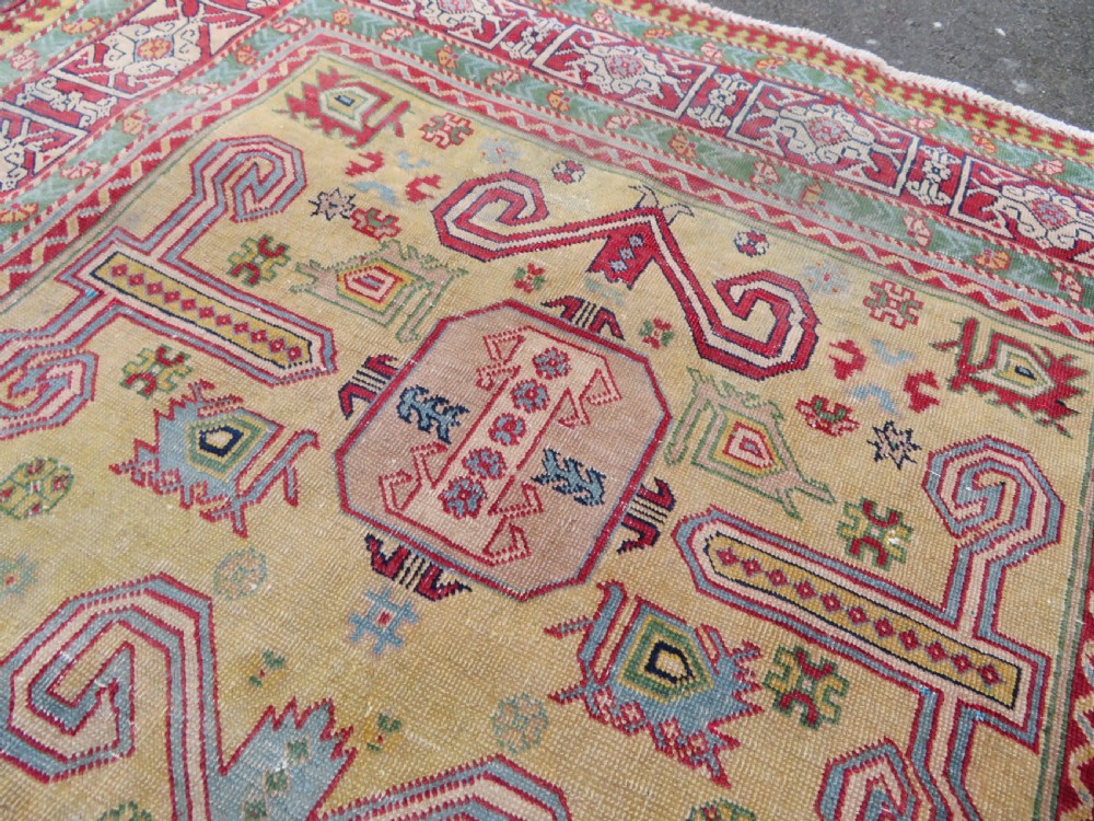 An antique hand-knotted Caucasian rug featuring a distressed yellow central field patterned with geometric ram horn motifs framed by red and green decorative borders. View XII