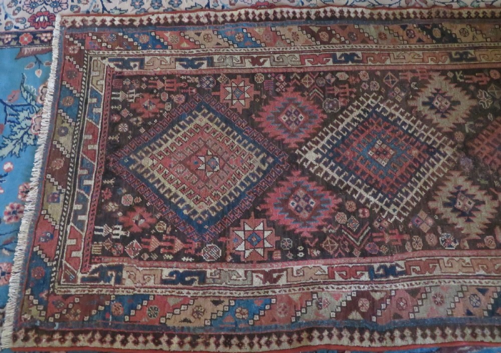 An antique hand-knotted wool runner rug featuring repeating geometric diamond medallions down a central dark field with multi-tiered tribal borders. View V