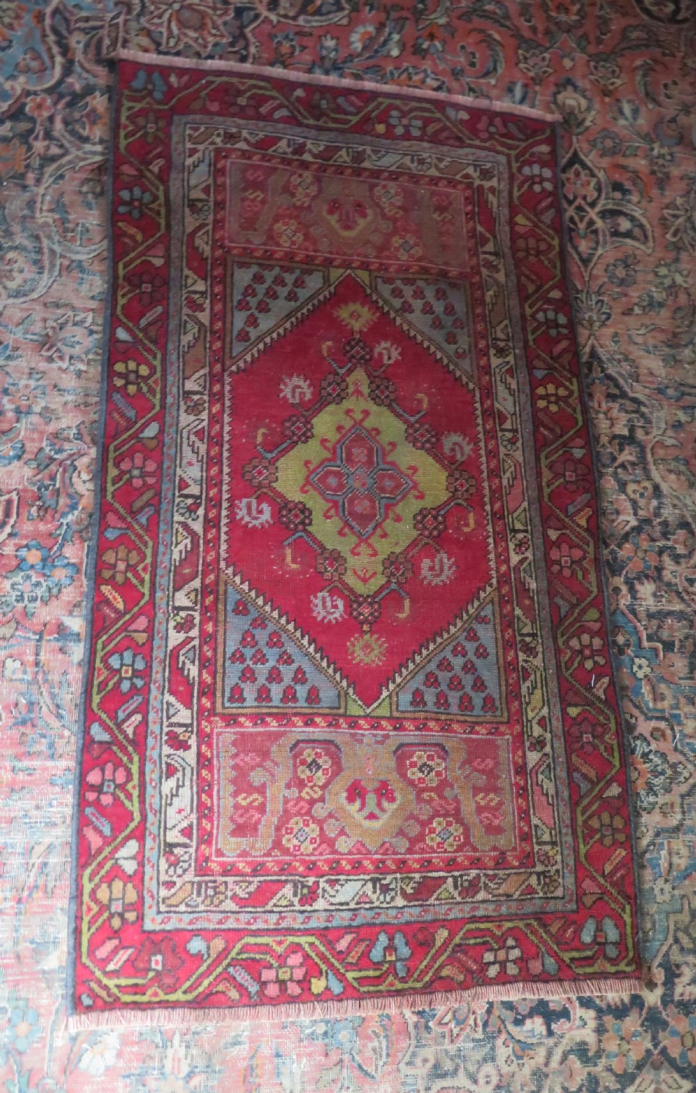 An antique hand-knotted Anatolian wool rug featuring a bold red field, a central geometric medallion in green and yellow, and detailed multi-patterned borders. View IV