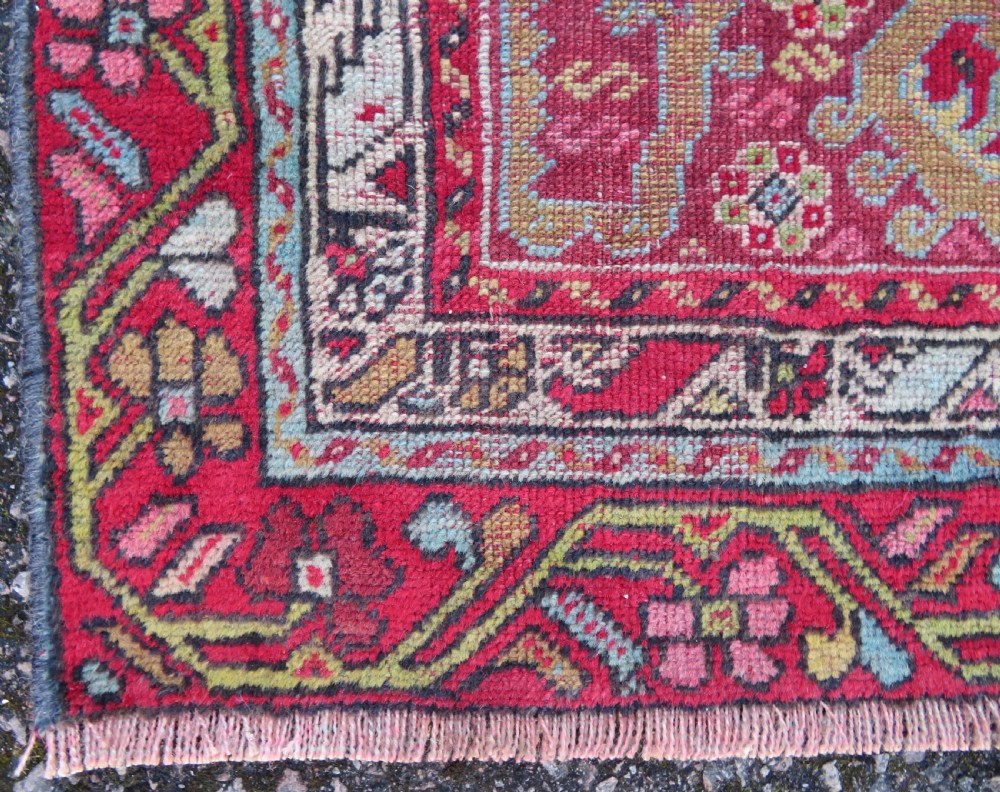 An antique hand-knotted Anatolian wool rug featuring a bold red field, a central geometric medallion in green and yellow, and detailed multi-patterned borders. View VI