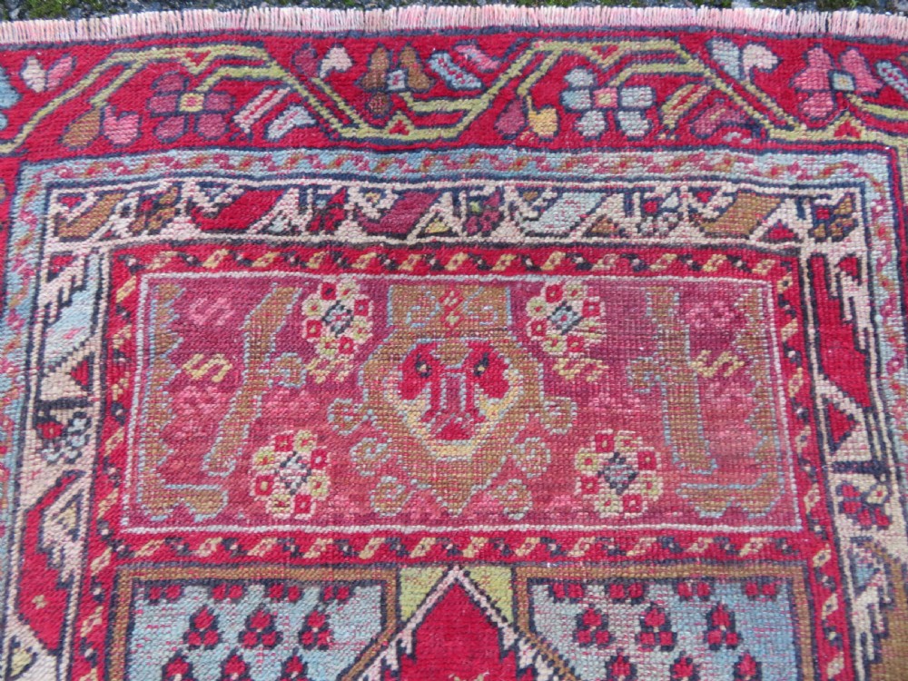 An antique hand-knotted Anatolian wool rug featuring a bold red field, a central geometric medallion in green and yellow, and detailed multi-patterned borders. View VII