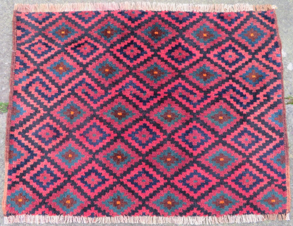 A square antique hand-knotted wool saddlebag face featuring a repeating geometric diamond pattern in red, navy blue, teal, and orange tones with light-coloured fringes.